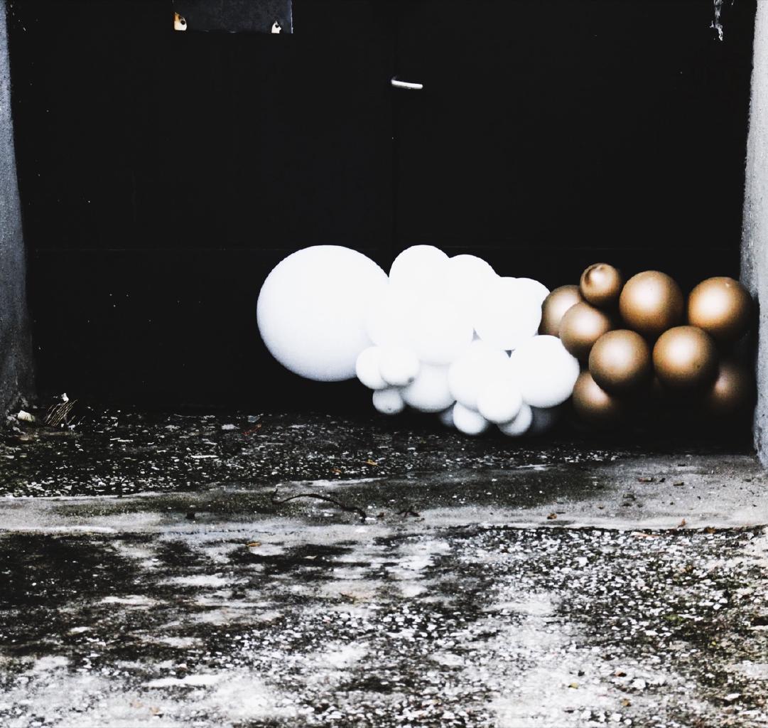 Σαν δύο ζώνες σκοτεινές, μια πάνω και μια κάτω, χωρισμένες απ’ αυτά τα μπαλόνια. Τι δουλειά έχουν μερικές νότες χαράς σ’ ένα πένθιμο εμβατήριο; Πως υπάρχουν μπαλόνια σ’ έναν διζωνικό ασφυκτικό χώρο; Η τέχνη έχει δώσει αναπαραστάσεις του ωραίου στις πιο αντίξοες γι’ αυτό (το ωραίο) να υπάρχει συνθήκες. Είναι σαν να χαμογελά ένα παιδί ενώ η πόλη του βομβαρδίζεται. Είναι όπως όταν βλέπεις μέσα απ’ το τσιμέντο να φύονται λουλούδια. Και είναι και μια προτροπή για τον άνθρωπο, κατά κανόνα σκοτεινή ύπαρξη που σώζεται απ’ τις φωτεινές εκλάμψεις της, να επιδαψιλεύει όπου μπορεί και λίγο θάμβος. Αυτή η πορεία απ’ την γέννηση ως τον θάνατο χρειάζεται παντού στασίδια για να ξεκουράζεται και να χαμογελά. Είναι στις αποζημιώσεις που δικαιώνεται το κόστος και η σπατάλη της ζωής.