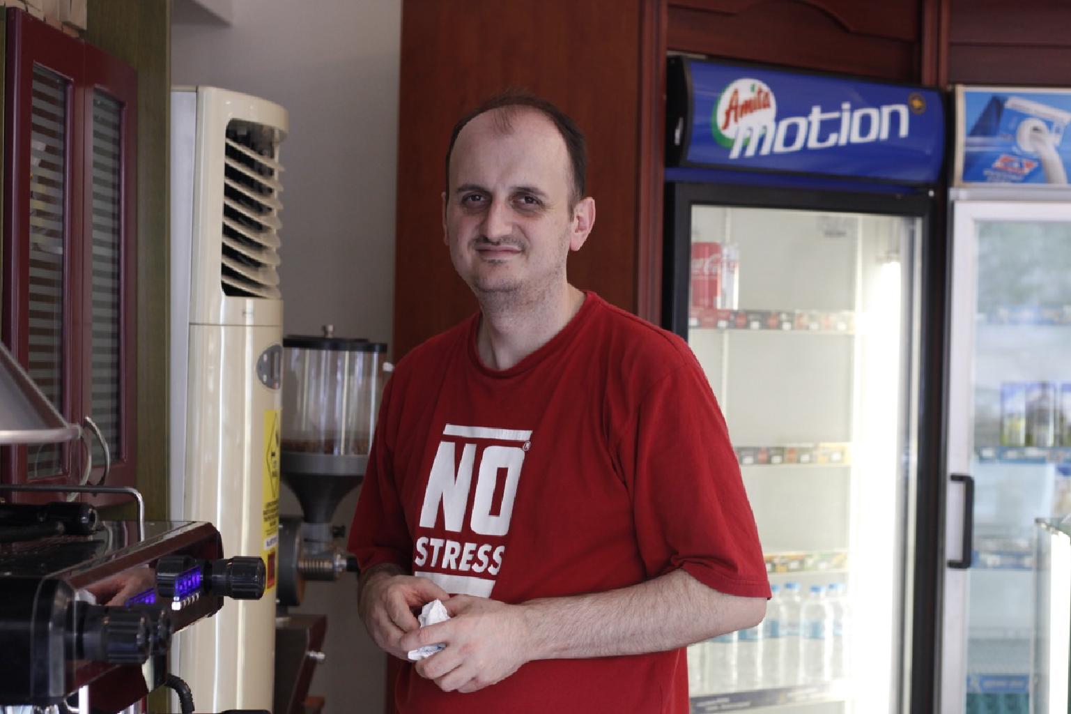 Πήρα δυνάμεις και βγήκα με φόρα στους δρόμους της Καλλιθέας εξοπλισμένος με τη φωτογραφική μου μηχανή. Άνοιξα ορίζοντες για να κινηθώ μες στην πυκνοκατοίκηση σαν σε ανοιχτωσιά. Βυθισμένος για τόσους μήνες στην κατάθλιψή μου, δεν ήξερα πως θα είχα και την έξοδο απ’ αυτήν. Με κυνήγησαν όλοι μανιωδώς. Κανείς δεν θέλει τον βαρύτιμο δίπλα του. Η αγάπη είναι για τον κολασμένο βασανιστήριο. Είμαι ο άγιος, ένας διαφορετικός άγιος, πονηρεμένος και συνάμα ψαρωμένος απ’ την ζωή. Πάναγνός και ευφυής. Φεύγω συνέχεια γιατι δεν θέλω να φθαρώ. Σταματάω λίγο στου Κώστα, είναι ο φούρναρής μου. 38 χρονών και ίσα χρόνια μόνο χαμόγελα. Νοιάζεται για όλους τους πελάτες του. Συναισθηματικά γενναιόδωρος με έχει περιμαζέψει στην μεγάλη του αγκαλιά. Χωράμε εκεί όλοι οι καταρρακωμένοι της ζωής. Ενδιαφέρεται για τις γευστικές μας μανίες και φτιάχνει στο ευρύχωρο αρτοποιείο του μια γωνιά με προϊόντα της αρεσκείας του καθενός μας. Θεαματικά πονόψυχος. Κι αυτή η γειτονιά μου, έχει, έχει τόσους αγίους, κάθε μέρα συναντιέμαι και μ’ έναν, σ’ αυτό το απάγκιο γωνία Αγίων Πάντων και Κρέμου που είναι ο Κώστας. Ο Κώστας μου μήνυσε τα νέα για τον θάνατό σου Αντώνη. Σε θυμάμαι γλυκό, με το σακάκι σου και την αύρα των δαιμόνων σου γύρω σου. Σε παρατηρούσα νομίζω για χρόνια. Είχες το φέγγος σχεδόν ανεπαίσθητου φωτοστέφανου. Δεν σε γνώριζα, μα όταν έμαθα για τον θάνατο σου ήταν σαν οι ιαματικές ματιές μου πάνω σου να μαζεύτηκαν όλες μαζί και να έγιναν μνήμες που μοιραστήκαμε μαζί. Καλό ταξίδι. (Στη φωτογραφία ο αρτοποιός μου Κώστας.)